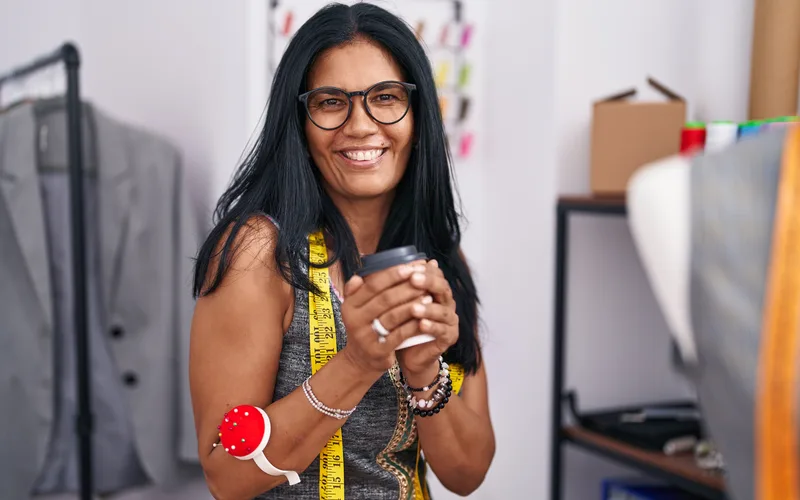 Costureira madura de óculos, com fita métrica no pescoço e alfineteiro no pulso, sorrindo com um café na mão no ateliê.