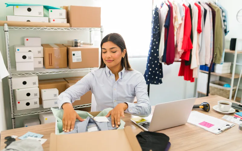 Dona de loja virtual embalando um pedido na mesa de trabalho, com notebook, estante de caixas e uma arara de roupas coloridas ao lado.