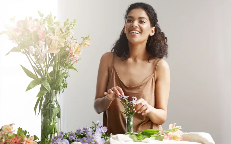Florista sorridente montando um buquê de flores delicadas numa bancada clara, com um vaso de lírios ao lado.