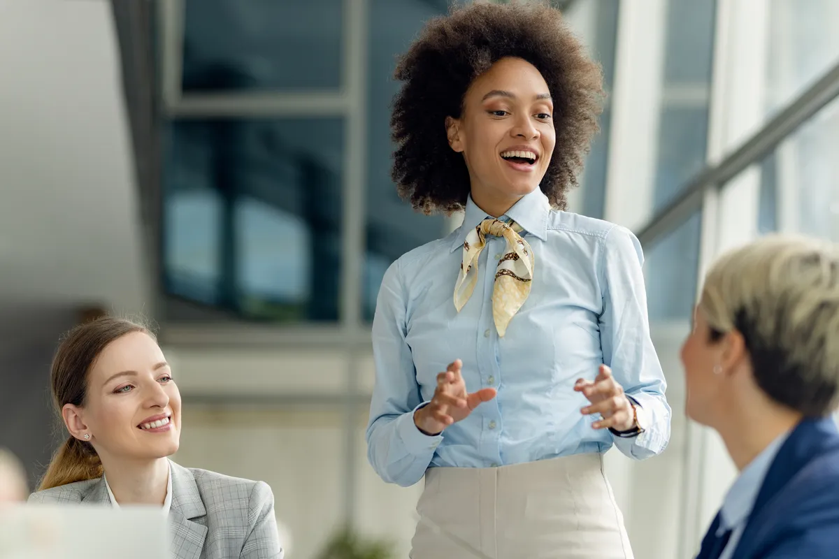 Líder de camisa clara e lenço no pescoço conversa animada com as colegas, que ouvem sorrindo, num escritório iluminado pela janela.