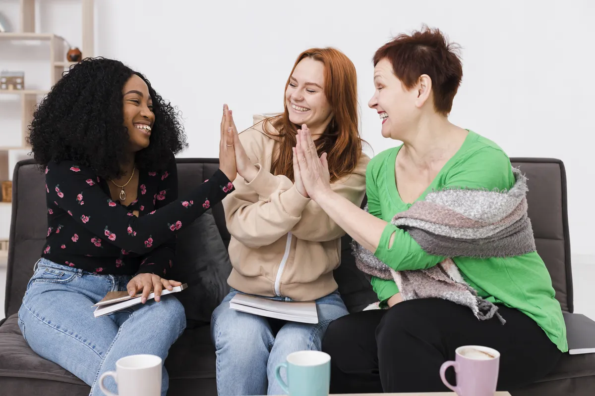 Três mulheres de idades diferentes sentadas lado a lado num sofá, rindo e batendo as mãos numa roda de conversa numa sala clara.